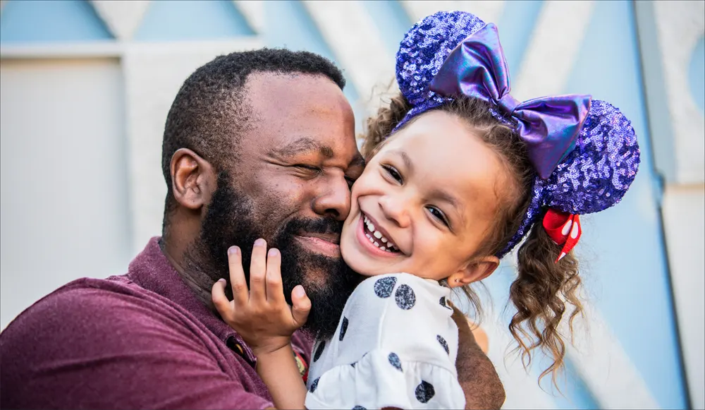 Disneyland dad hugs his little girl who has a purple Minnie Mouse hat on her head.